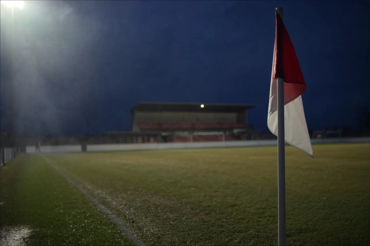 A floodlit suburban football ground at dusk, corner flag in the foreground and an empty stand beyond