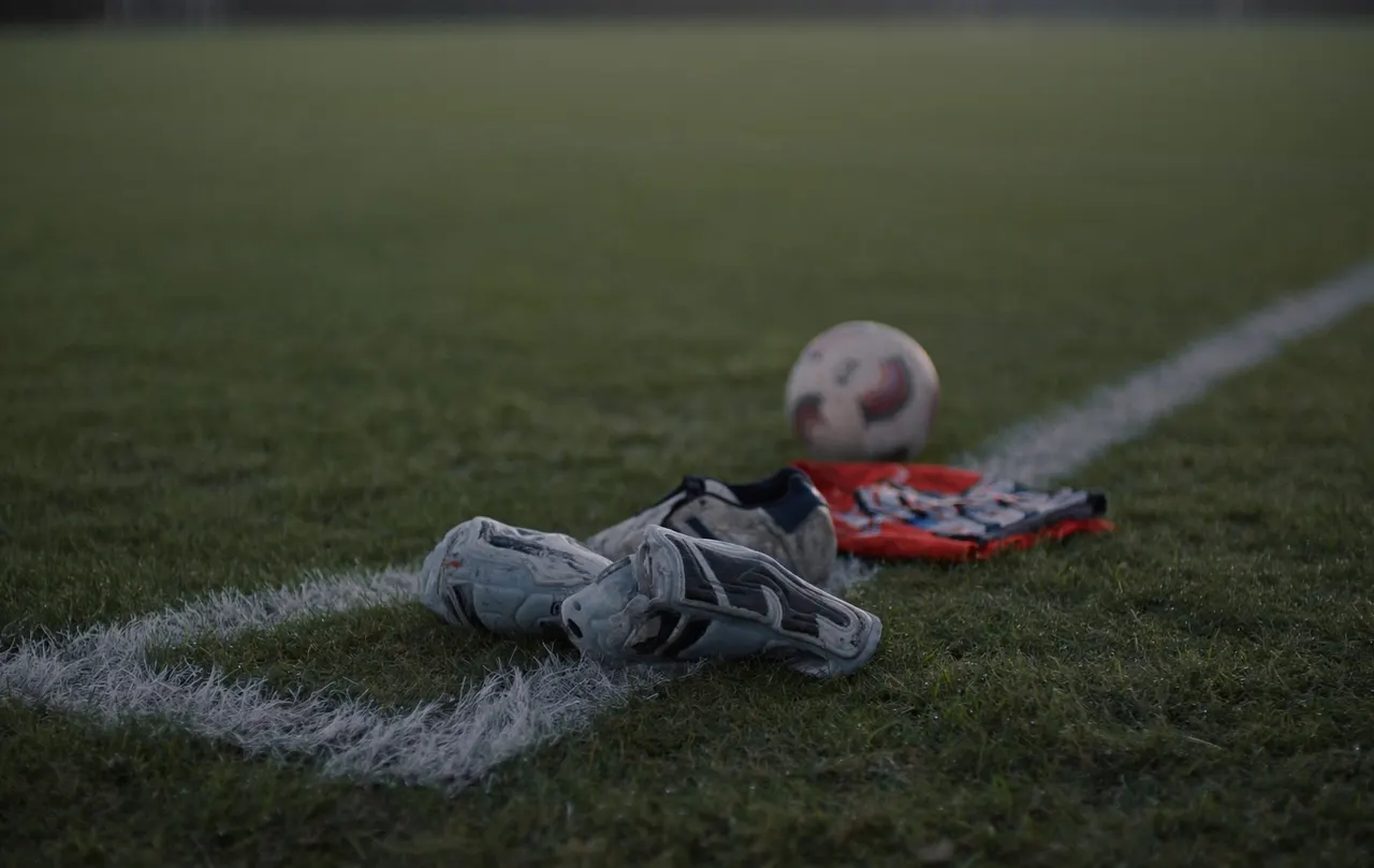 Football boots, shin pads, a match ball and a folded training bib laid out on the touchline of a dewy grass pitch